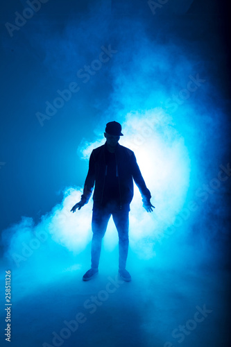 Silhouette of man giving solo performance, dancing alone in hip hop style on club scene with blue neon lightning and smoke.
