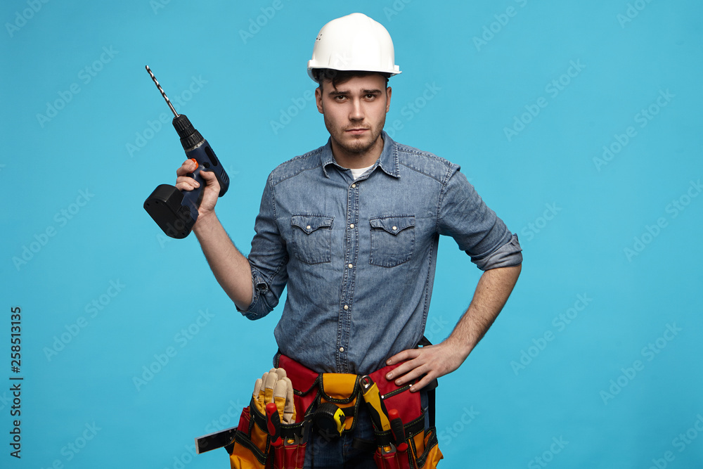 People, repair, equipment and renovation concept. Stylish young Caucasian repairman wearing hardhat, tool belt and overalls going to hang picture on wall, posing in studio with drill in his hand