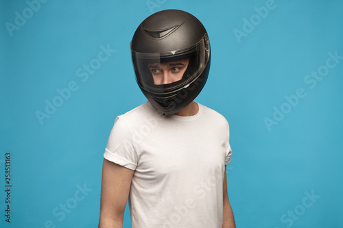 Portrait of stylish young man wearing white t-shirt and motorcycle helmet posing isolated at blue studio wall looking away. Motor sports, extreme lifestyle, transportation and people concept © Anatoliy Karlyuk