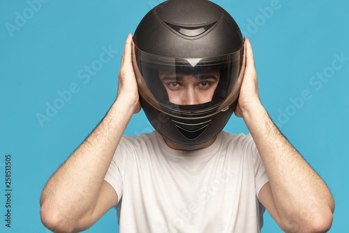 Isolated studio shot of self determined serious young Caucasian male biker taking on stylish black motorcycle helmet, going to have ride on his motorcycle staring at camera with confident look © Anatoliy Karlyuk