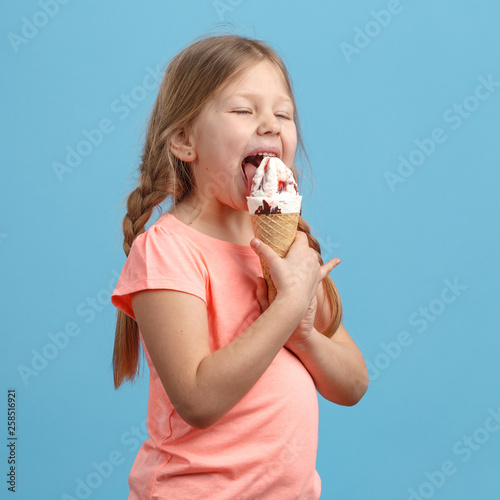 Cute little girl eats ice cream and laughs in studio over blue background