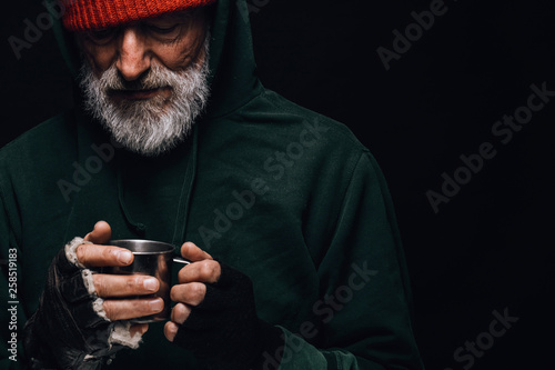 Old homeless man with grey beard covering up in green decrepit wear holding a mug of hot tea to warm himself in a cold night photo