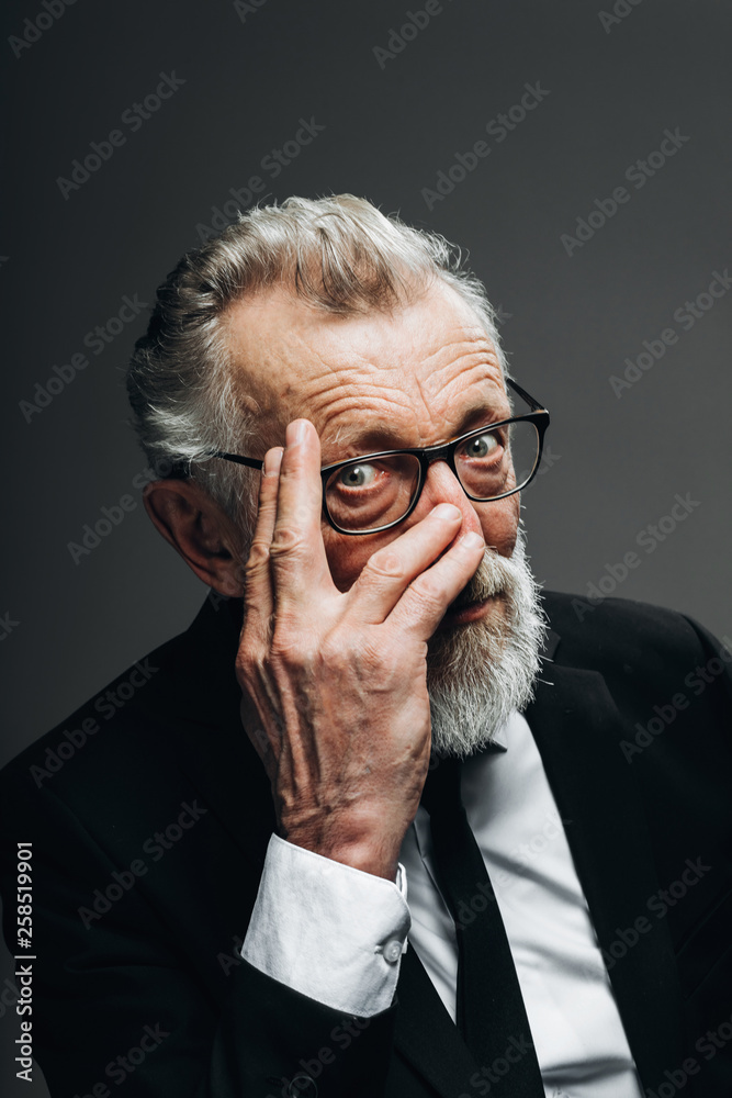 Mature bearded politician in eyewear and confident experienced eyes, dressed in black formal suit thinking with hand under chin posing isolated over black Mature bearded politician in eyewear and confident experienced eyes, dressed in black formal suit thinking with hand under chin posing isolated over black