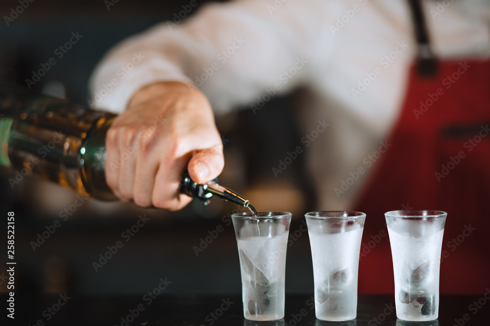 Close up photo of hands of a bartender pouring some drink from the bottle into shot frozen glasses on a wooden counter. Close up photo of hands of a bartender pouring some drink from the bottle into shot frozen glasses on a wooden counter.