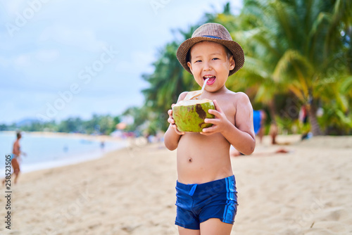 Tanned Asian boy stands on the beach in a hat and drinks coconut