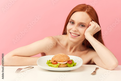 Excited red-haired skinny half naked woman in underwear, with greedy eyes being ready to eat burger, sitting at table isolated over pink background. Eating disorder.