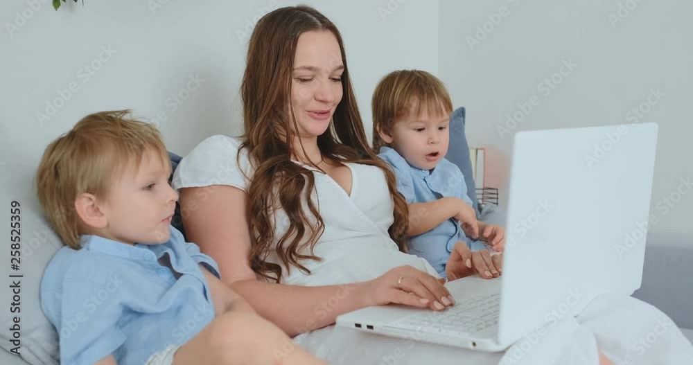 Beautiful young mom and two little kids boys are looking at the laptop screen family photos. And they do online shopping. Call grandmother through video chat. Children show their fingers on the laptop Beautiful young mom and two little kids boys are looking at the laptop screen family photos. And they do online shopping. Call grandmother through video chat. Children show their fingers on the laptop