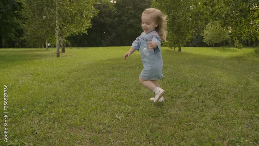 Baby girl running through the grass and catching soap bubbles in garden in sunset sunlight. Slow motion. Happy smiling baby girl. Baby girl running through the grass and catching soap bubbles in garden in sunset sunlight. Slow motion. Happy smiling baby girl.