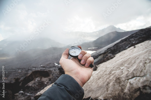 A first-person view of a man's hand holding a magnetic compass against a background of Caucasus mountains and a cracked glacier dusted with volcanic dust. The concept of traveling