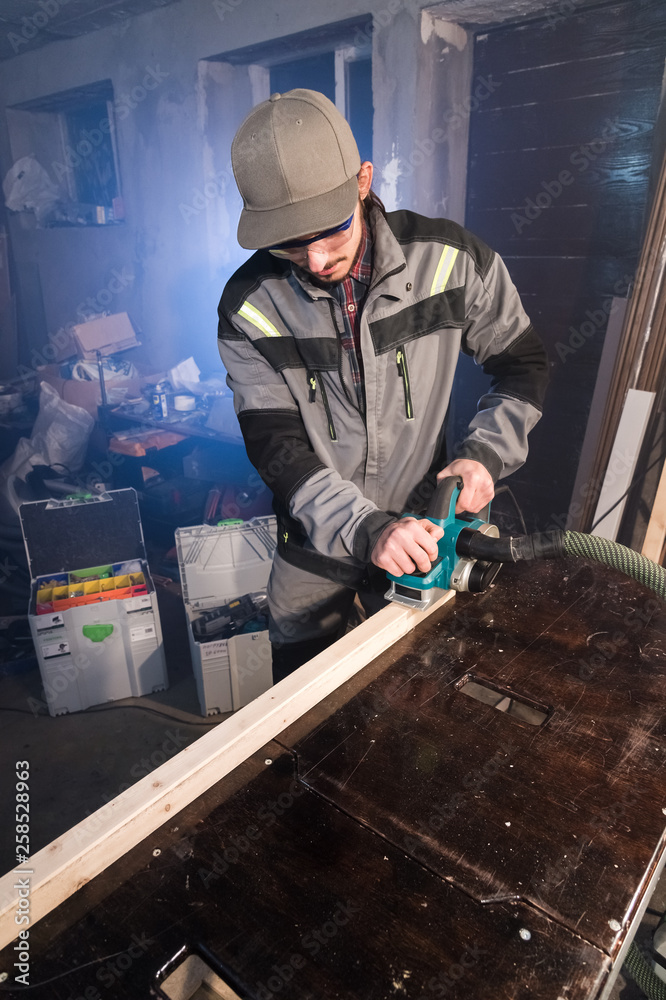 Portrait of a young carpenter working with an electrical plane in a home wood workshop. The concept of a business idea and a woodworking startup. Wide angle