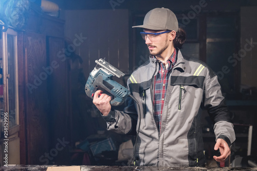 Portrait of a young carpenter joiner in overalls wearing a cap and goggles with an electric jigsaw in hand at a home workshop. Starting a business. Craftsman