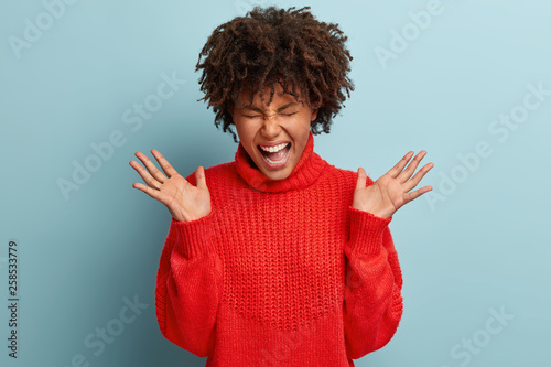 Positive curly ethnic woman gestures actively, spreads palms, shouts emotionally, rejoices coming wedding day with boyfriend, wears red casual jumper, closes eyes, isolated over blue background