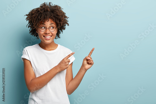 Studio shot of optimistic Afro American woman with crisp hair, positive smile, points aside with fore fingers, wears round spectacles and white t shirt, shows awesome thing at upper right corner photo