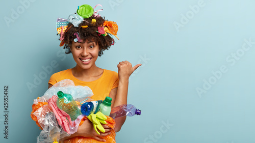 Cheerful ethnic woman with curly hair, carries trash, points away with thumb, picks up litter, reduces plastic wastes cares of envirnoment stands over blue background involved in campaing for cleaning photo