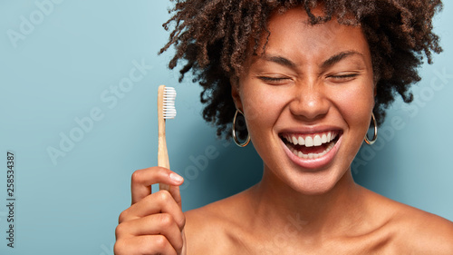 Dental health concept. Happy smiling dark skinned woman with curly hair, laughs while has morning routines, shows bright smile, holds toothbrush, stands with bare shoulders over blue background