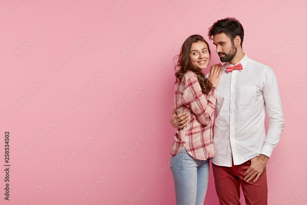 Horizontal shot of happy romatic couple embrace and have pleasant talk, wear stylish elegant clothes, smile happily pose over pink background with free space left for your advertisement or information Horizontal shot of happy romatic couple embrace and have pleasant talk, wear stylish elegant clothes, smile happily pose over pink background with free space left for your advertisement or information