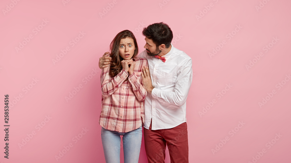 Stupefied female stares with bugged eyes, keeps hands together, her boyfriend embraces and asks not be afraid, dressed in elegant clothing, isolated over pink background. People and relationship Stupefied female stares with bugged eyes, keeps hands together, her boyfriend embraces and asks not be afraid, dressed in elegant clothing, isolated over pink background. People and relationship