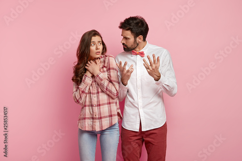 Angry man reproaches girlfriend, gestures and explains something angrily, frustrates puzzled woman keeps hands together, afraids of husbands remarks, sort out relationship, isolated on pink wall