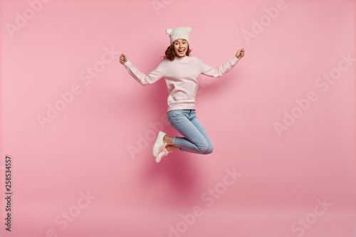 Full length body shot of joyful pleased young female model jumps happily in air against pink background, wears warm hat with ears, sweatshirt, jeans and sneakers, feels energetic and optimistic