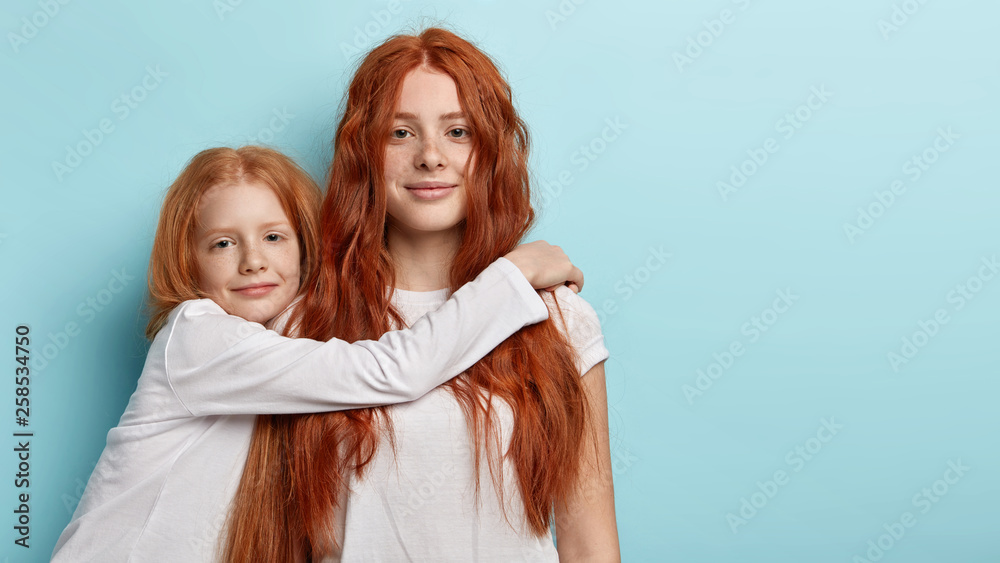 Two adorable foxy girls sisters embrace indoor, look at camera with green eyes. Pretty freckled little child embraces with love best friend, have friendly relationships. I am always by your side - obrazy, fototapety, plakaty 