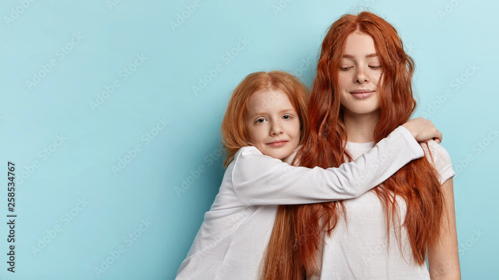 Horizontal shot of small girl embraces older sister with love, missed her very much, wants to play together, glad to have each other, look alike, have truthful friendship. Family, relationship concept Horizontal shot of small girl embraces older sister with love, missed her very much, wants to play together, glad to have each other, look alike, have truthful friendship. Family, relationship concept