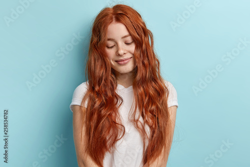Indoor shot of timid ginger teenage girl looks down with dreamy pleased thoughtful expression and tender smile has long wavy foxy hair, dressed in casual white t shirt, isolated on blue background