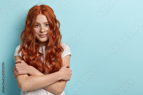 Teens and beauty concept. Pretty teenager with healthy freckled skin, no make up, wears white t shirt, embraces herself, has timid facial expression, feels shy during first date, stands over blue wall