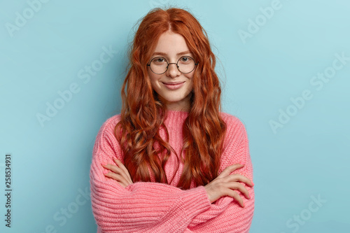 Satisfied ginger charming woman with curly hair, wears transparent glasses, keeps arms folded over chest, has pleasant talk with contemporary, wears oversized clothes, isolated over blue background photo