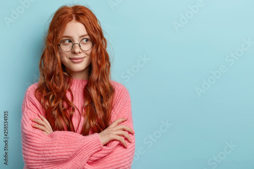 Φωτογραφία Pensive dreamy lovely young ginger female with pleasant appearance, imagines dreams come true, keeps hands crossed, dressed in oversized jumper, wears optical glasses, isolated over blue wall