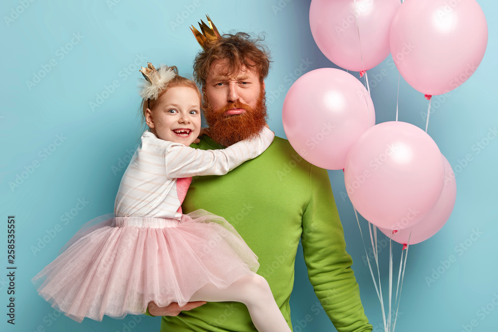 Family celebration concept. Tired ginger dad holds small female kid, feels fatigue after amusing children on party, pose near air balloons against blue background. Time for fun during holiday Family celebration concept. Tired ginger dad holds small female kid, feels fatigue after amusing children on party, pose near air balloons against blue background. Time for fun during holiday