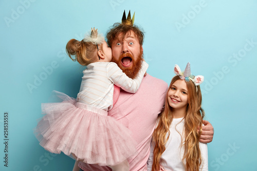 Mad father hears secret from daughter, spends free time with children, embraces and shares love, wear carnival costumes for party, isolated over blue wall. Kids and parents concept. Festive event
