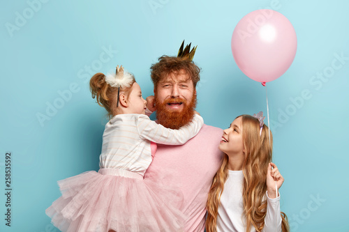 Billede på lærred Fatigue depressed red entertainer has crown on head, looks at camera with miserable expression, surrounded with kids, organizes noisy birthday party, pose over blue background