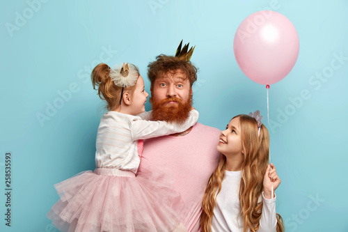 Busy father organizes holiday on International childrens day for two daughters, have home party, wear crowns and festive clothes. Little adorable girl holds air balloon, looks at dad and sister