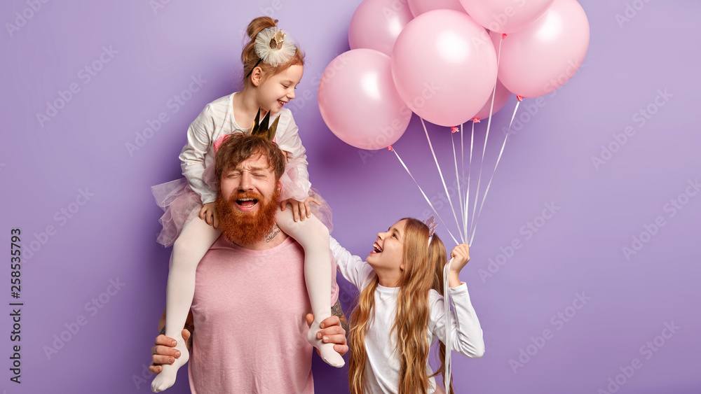 Fototapeta premium Puzzled tired busy father entertains daughters, feels fatigue of playing different games on party, enjoy birthday celebration stands against purple background. Funny sisters look happily at each other