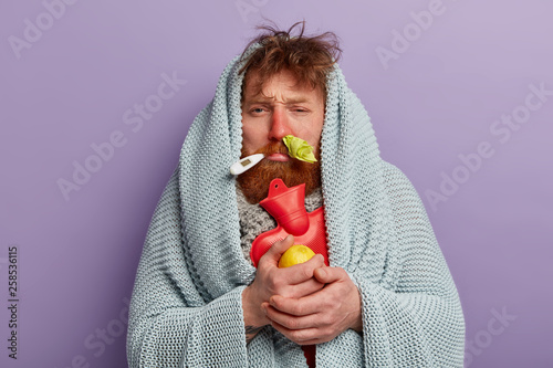 Photo of upset sick man has red nose from rhinitis, trembles under coverlet, warms with hot water bag, has thermometer in mouth, holds fresh lemon, isolated on lilac wall. Health and personal care