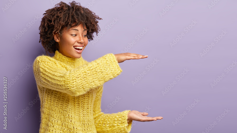 Sideways shot of glad dark skinned lady shapes big object with both hands, has sincere smile, being in good mood, shows which huge present she got, wears knitted yellow jumper, stands indoor Sideways shot of glad dark skinned lady shapes big object with both hands, has sincere smile, being in good mood, shows which huge present she got, wears knitted yellow jumper, stands indoor