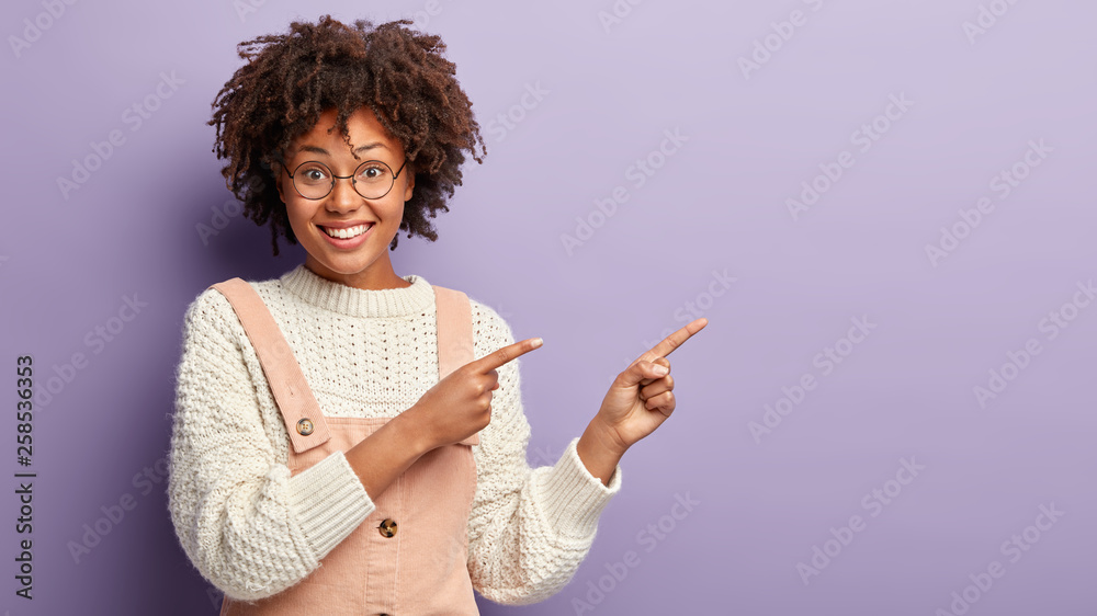 Pleased Afro American lady points aside at blank space, suggests to have look, shows direction, smiles happily, wears white jumper, pink overalls isolated over lilac studio wall. Advertisement concept Pleased Afro American lady points aside at blank space, suggests to have look, shows direction, smiles happily, wears white jumper, pink overalls isolated over lilac studio wall. Advertisement concept
