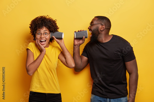 Joyful dark skinned couple have fun drinking coffee, hold paper cup near ear and mouth. Students have spare time after classes, drink hot beverage, wear black and yellow t shirts, pose indoor
