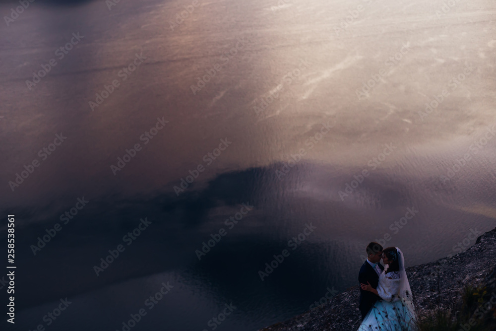 river water with reflection. Newlyweds hug on the rocky shore of