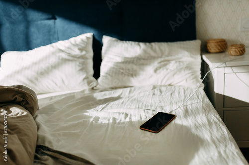 Morning light on the pillows, empty bed and unmade at the first light of dawn, battery mobile phone charging and lying on white sheet