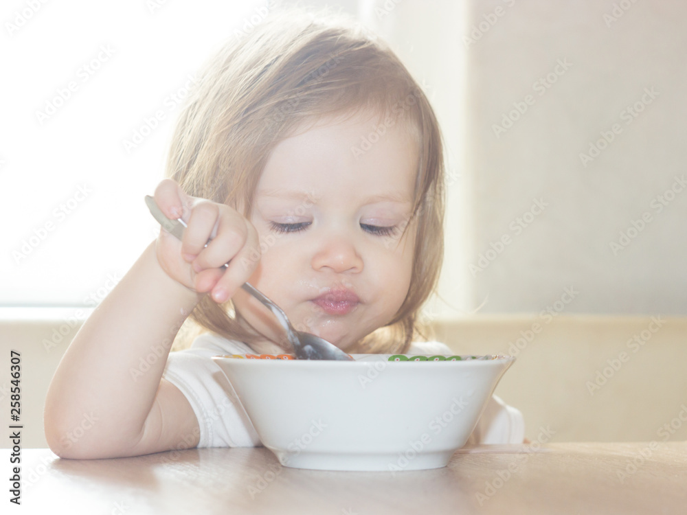 Little shaggy baby girl with pleasure alone holds a spoon and eats delicious food from a white plate.