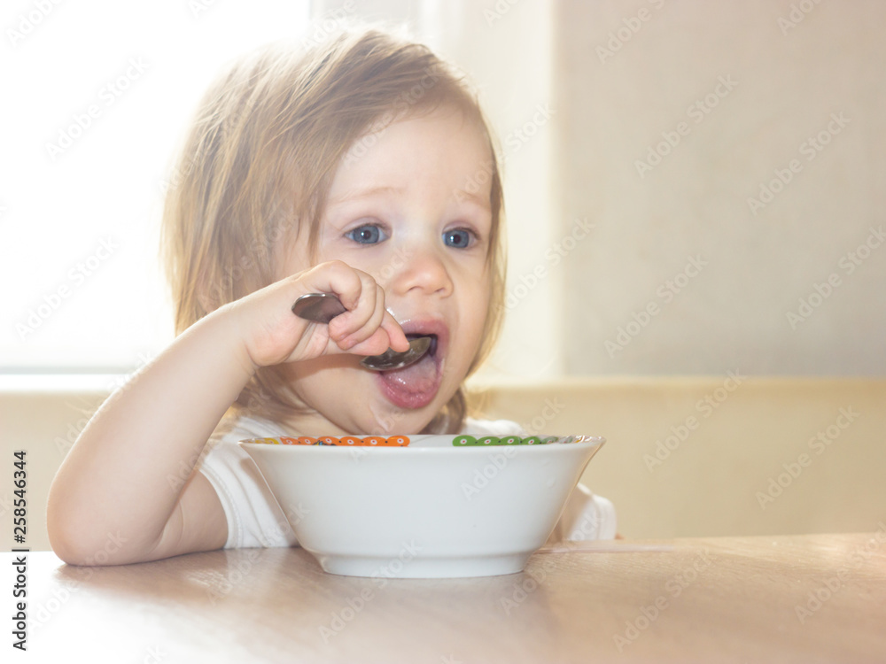 Little shaggy baby girl with pleasure alone holds a spoon and eats delicious food from a white plate. Little shaggy baby girl with pleasure alone holds a spoon and eats delicious food from a white plate.