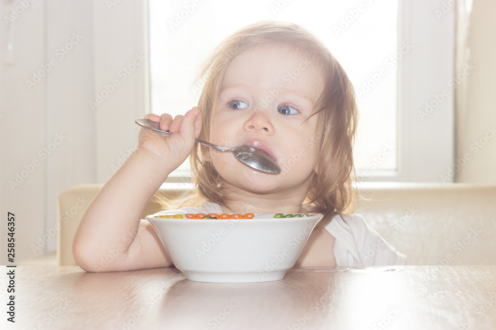 Little shaggy baby girl with pleasure alone holds a spoon and eats delicious food from a white plate. Little shaggy baby girl with pleasure alone holds a spoon and eats delicious food from a white plate.
