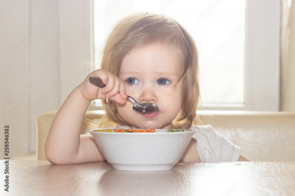 Little shaggy baby girl with pleasure alone holds a spoon and eats delicious food from a white plate. Little shaggy baby girl with pleasure alone holds a spoon and eats delicious food from a white plate.
