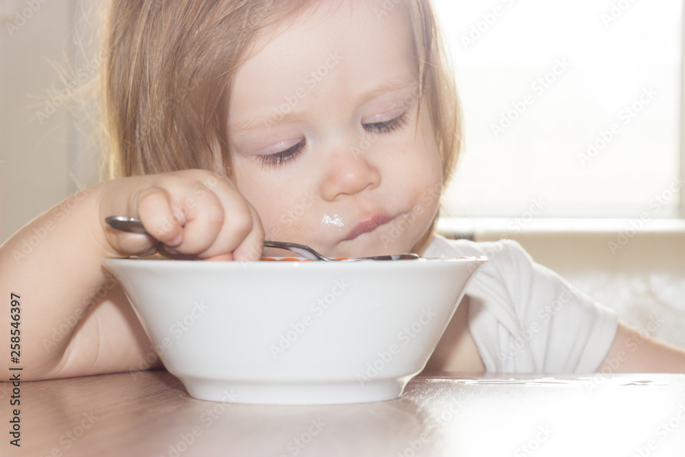 Little shaggy baby girl with pleasure alone holds a spoon and eats delicious food from a white plate. Little shaggy baby girl with pleasure alone holds a spoon and eats delicious food from a white plate.