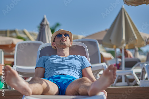Cute man in sunglasses and sunhat is laying in the sunbed and enjoying his rest in the hotel.