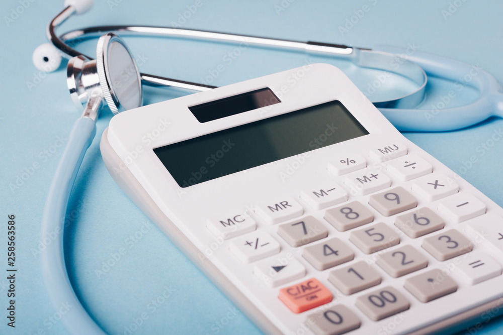 White calculator and blue stethoscope on a blue background as a symbol of paid (expensive) medicine White calculator and blue stethoscope on a blue background as a symbol of paid (expensive) medicine