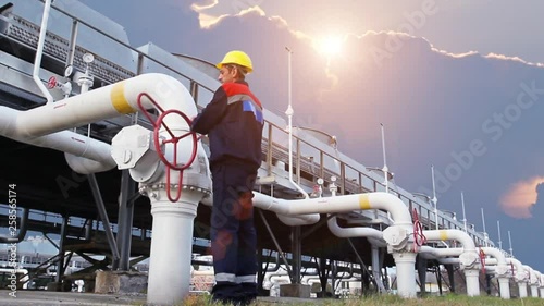 worker closes ball valve on cooling installations at gas compressor station, against background of sunset