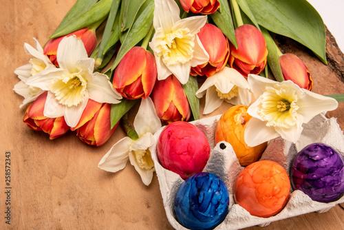 Bunte Blumen Tulpen und Narzissen sowie gefärbte Ostereier auf einem Holzbrett