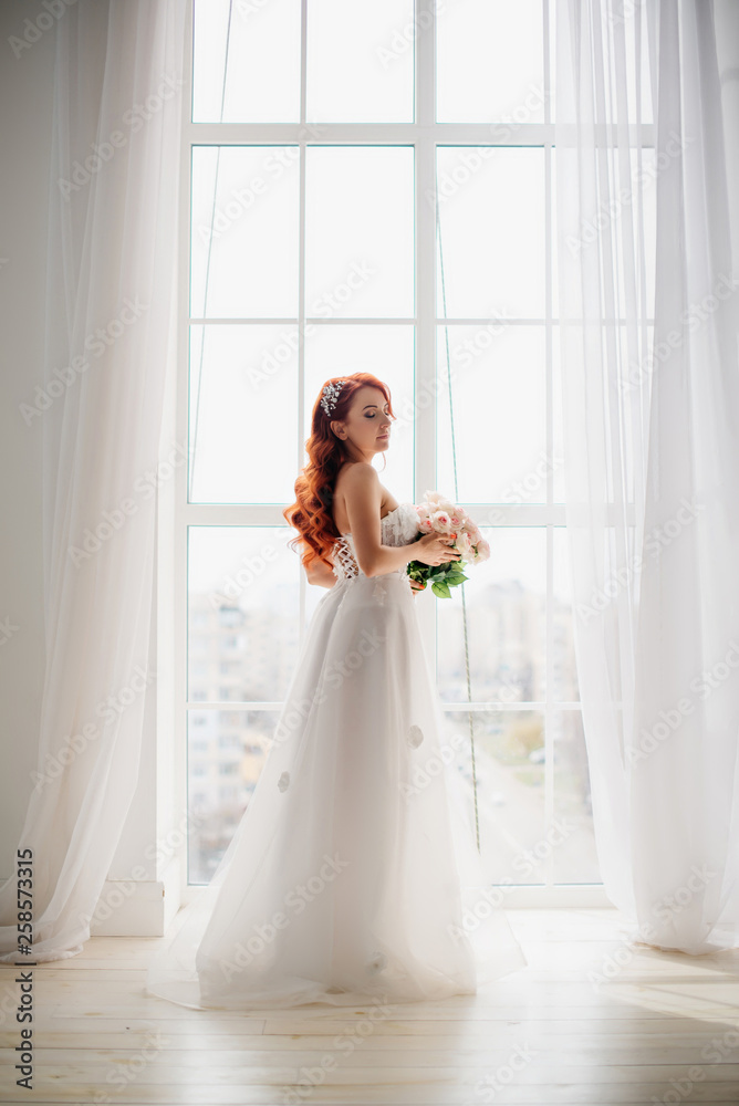 Red-haired bride in white wedding dress stands near large window Red-haired bride in white wedding dress stands near large window